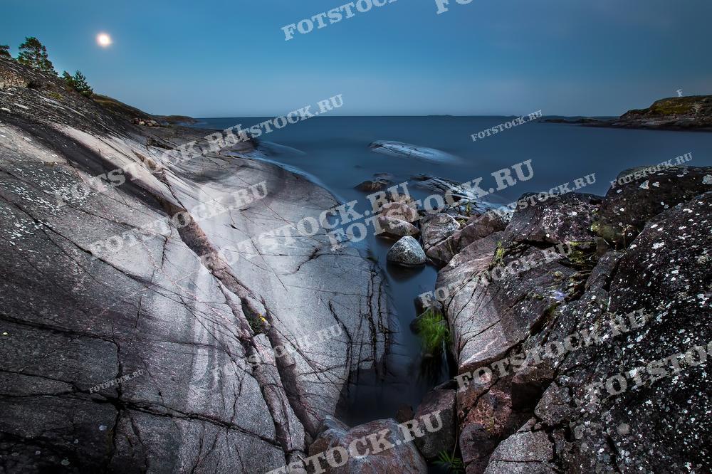 На фотографии изображен берег Ладожского озера в Карелии, освещенный лунным светом. Гладкие, серо-розовые скалы создают впечатление величественного и древнего пейзажа. Спокойная вода озера отражает лунный свет, усиливая атмосферу тишины и умиротворения. Это место привлекает своей природной красотой и возможностью насладиться уединением в ночное время.
